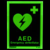 a front shot of the aed location sign glowing in the dark with text stating AED Emergency Defibrillator and a green heart with a lightening bolt through it with a first aid cross in the top right corner