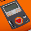 Close-up of an AED control panel with a digital display, two orange-bordered buttons, and icons for battery status, maintenance, and electrode pads.